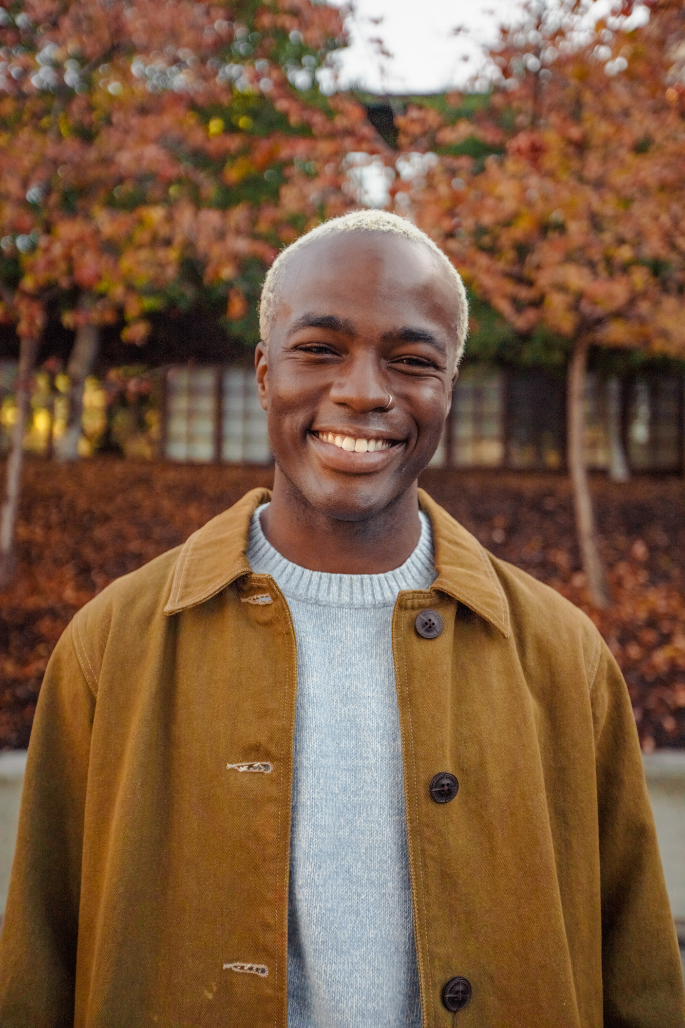 Photo of a Black man with blonde hair smiling and wearing a brown coat