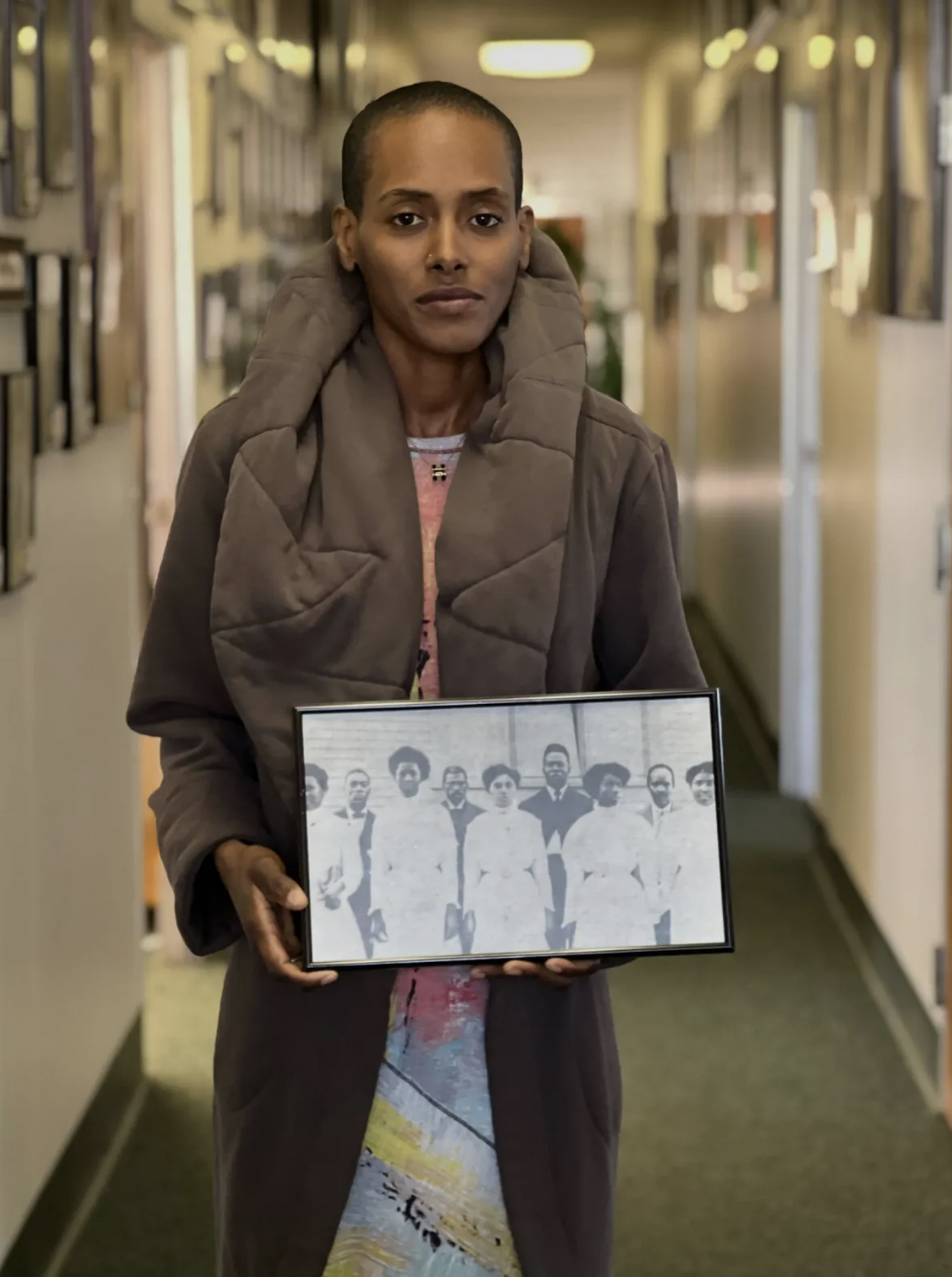 a photo of a Black woman looking straight at the camera. She is holding an old black and white photo with depicting nine Black people standing in front of a church.